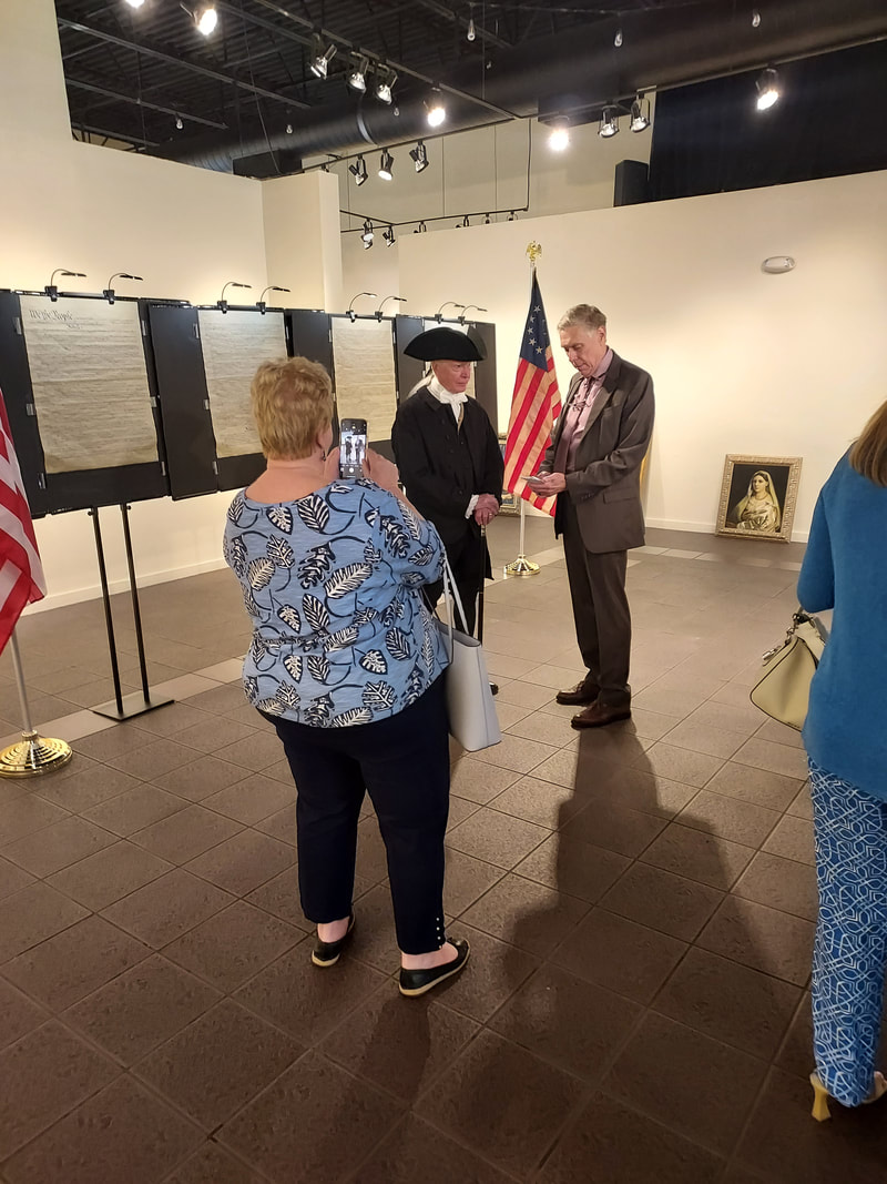 Costumed historical interpreter speaks with a man in a suit at an exhibition, while a visitor takes a photo. U.S. flag and documents displayed prominently.