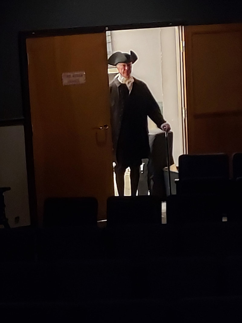 A man dressed in 18th-century clothing, including a large hat, stands at a partially open door, holding a cane, in a dimly lit theater setting.