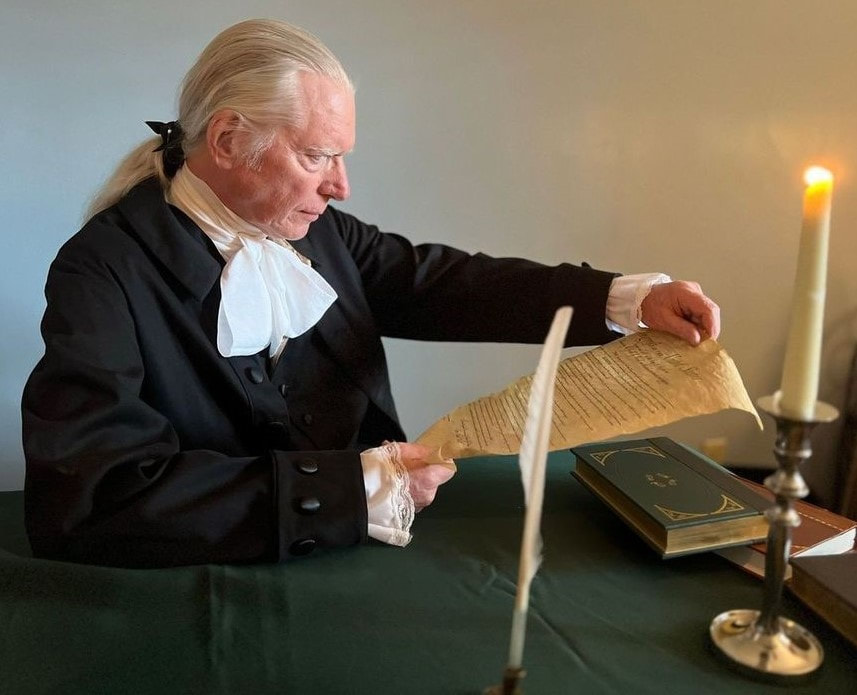 Historically dressed man examines an ancient document at a table, with a candle and books, highlighting a moment of research or reflection.