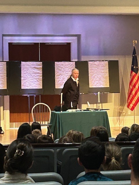 A speaker stands at a table during a presentation in a theater, with an American flag visible and an engaged audience in the foreground.