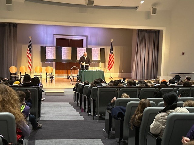 A speaker stands at a table in front of an audience, with U.S. flags flanking the stage. The event appears educational, engaging attendees in a presentation.