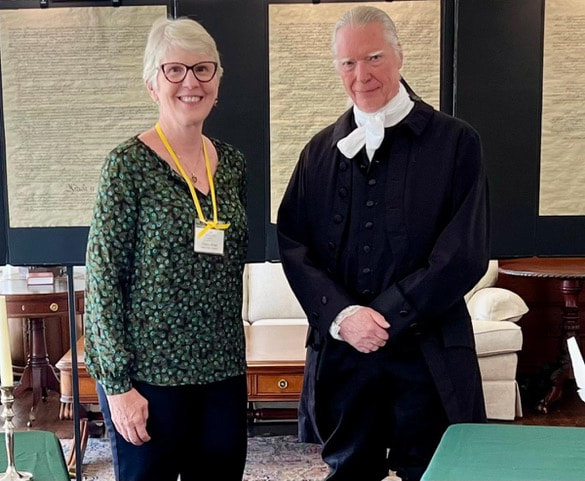 A woman in a patterned top stands next to a man in colonial attire, with historical documents displayed in the background, highlighting a cultural event.