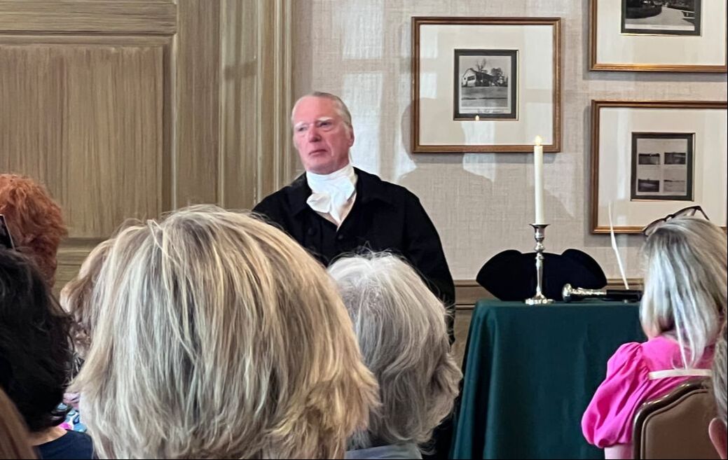 A speaker in historical attire addresses an audience, surrounded by framed photos and a candle, highlighting a cultural or educational event.